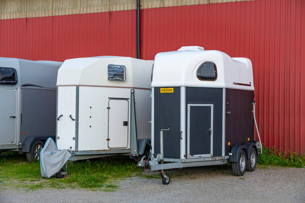 Reboques modernos de transporte de cavalos estacionados na grama perto de galpão de hangar sueco vermelho. - Foto, Imagem