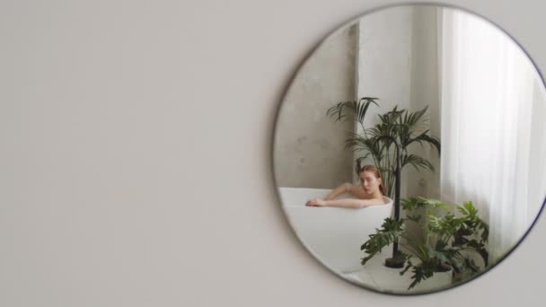 Zrcadlový odraz mladé blondýny ležící v moderní volně stojící vaně a pózující pro kameru - Záběry, video