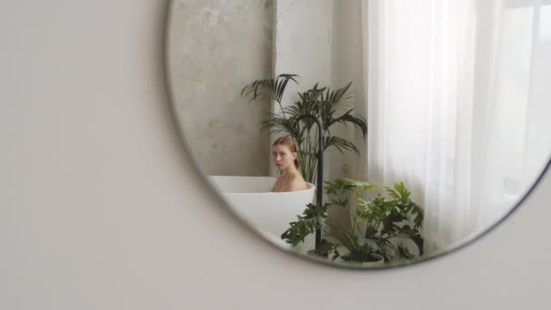 Riflessione nello specchio della giovane bella donna seduta nella moderna vasca da bagno indipendente e guardando la fotocamera - Filmati, video