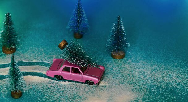 Malé růžové auto nese vánoční stromek na střeše zasněženým lesem hraček - Fotografie, Obrázek
