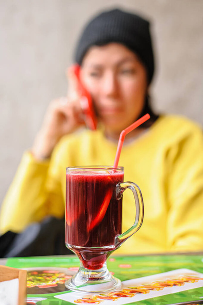 γυάλινο κύπελλο με ζεστό κρασί που περιλαμβάνει senby κόκκινο κρασί και φέτες πορτοκάλι με καλαμάκι στέκεται σε ένα τραπέζι σε ένα καφέ με φόντο μια νεαρή γυναίκα που μιλάει στο τηλέφωνο - Φωτογραφία, εικόνα