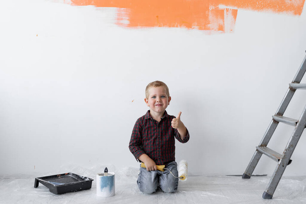 Ένα νεαρό αγόρι με χαμόγελο κάθεται σε ένα δωμάτιο κοντά στις σκάλες. Κρατάει ένα ρολό μπογιάς στα χέρια του με τον αντίχειρα ψηλά. Σχέδιο ανακαίνισης σπιτιού. - Φωτογραφία, εικόνα