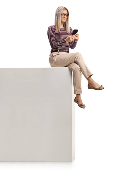 Woman with glasses using a smartphone and sitting on a tall white wall isolated on white background - Valokuva, kuva