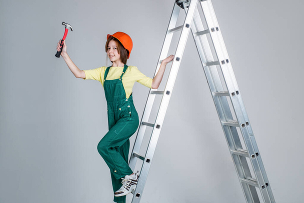 girl child wearing uniform and helmet while working in workshop with hammer on ladder, career - Foto, imagen