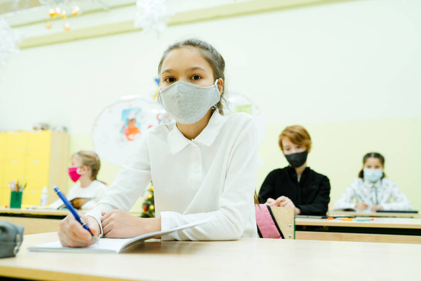 A girl in a protective mask sits at a desk and writes notes in a notebook - Photo, Image