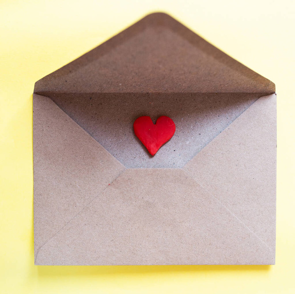Love letter in a craft envelope with clay red heart on yellow background. - Fotó, kép