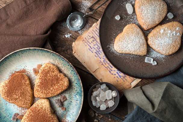 Herzförmige Kekse werden mit Kakao und Puderzucker auf einem Vintage-Hintergrund bestreut. Der Blick von oben. Das Konzept der Desserts. - Foto, Bild