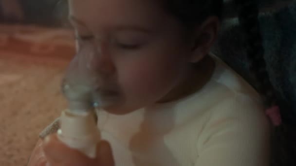 Klein meisje maakt inhalatie met behulp van een compressor inhalator terwijl ze op een bank zit. Vijf jaar geleden. meisje inhaleert zoutdamp met vernevelaar masker op haar gezicht. Moeder helpt haar dochter bij het inademen thuis - Video