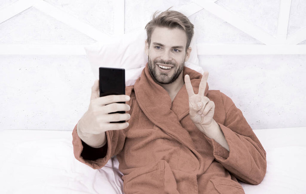 Selfie star. Happy man taking selfie with smartphone in bed. Handsome guy smiling with V hand gesture to selfie camera in mobile phone. Enjoying selfie session from bedroom - Photo, image