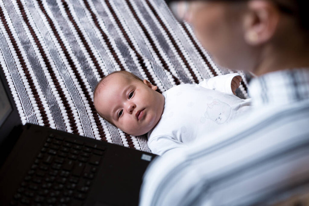 Παιδική ηλικία, μητρότητα, οικογενειακές έννοιες - επιτυχημένη χαμογελαστή πλούσια επιχειρηματίας Γυναίκα μητέρα κάθεται στο κρεβάτι με βρέφος το μωρό μιλάμε στο τηλέφωνο εργασίας σε φορητό υπολογιστή στο σπίτι σε απόσταση σε εσωτερικούς χώρους - Φωτογραφία, εικόνα
