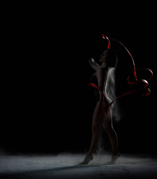 beautiful caucasian woman in a black bodysuit with a sports figure dancing with red ribbon on a black background, particles of flour fly in different directions - Zdjęcie, obraz