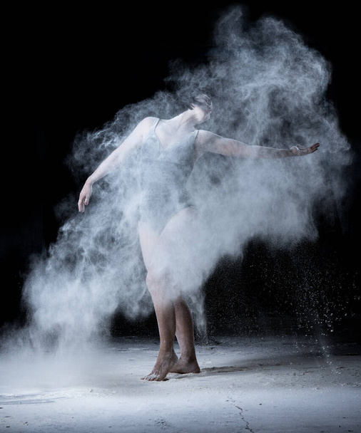 mulher com uma figura esportiva dançando em uma nuvem de farinha branca espalhada em um fundo preto, a dançarina está vestida com um bodysuit preto - Foto, Imagem