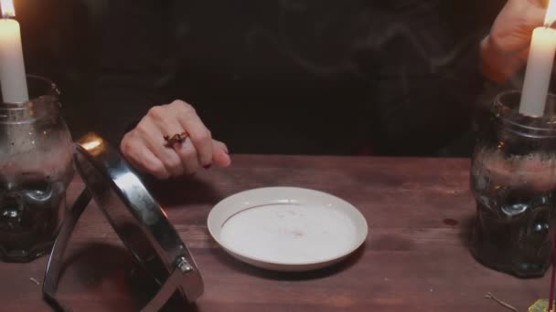 Senior donna strega indovino tiene specchio in una mano e accende candela rossa e cera gocciolante in un piatto in terribile rituale magico - Filmati, video