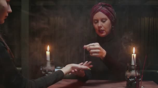 Mujer anciana adivina usa aguja con hilo para decirle el futuro a su joven cliente - Metraje, vídeo