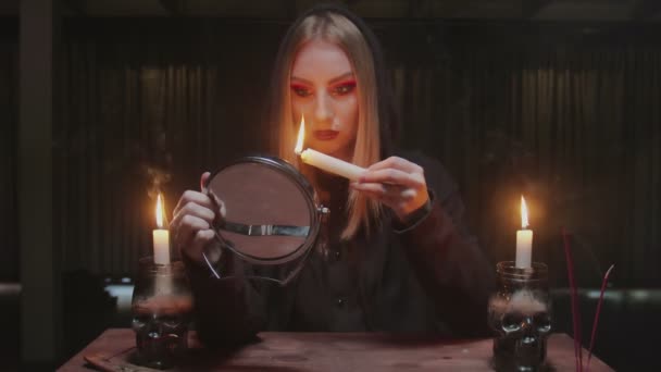 Joven bruja adivina sostiene vela en uno y mira al espejo en terrible ritual mágico - Metraje, vídeo