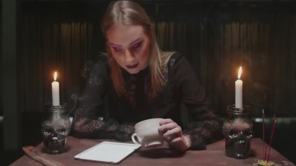 Молодая ведьма-гадалка держит чашку кофе и смотрит на табличку с предсказанием судьбы - Кадры, видео
