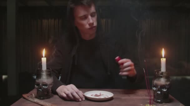 Concentrated mysterious wizard male fortune teller lights a red candle, hard puts it on plate in magic ritual - Footage, Video