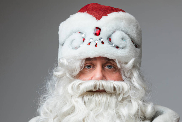 Headshot of Santa Claus in red hat, gray-haired beared tired man on gray studio background - Фото, зображення