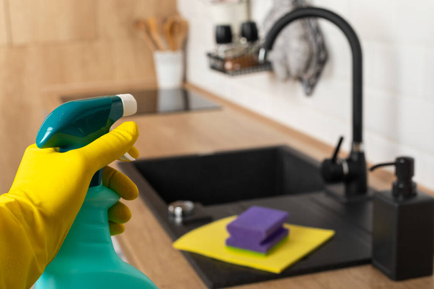 Housekeepers hand holding cleaning supplies in front of black kitchen sink - Φωτογραφία, εικόνα
