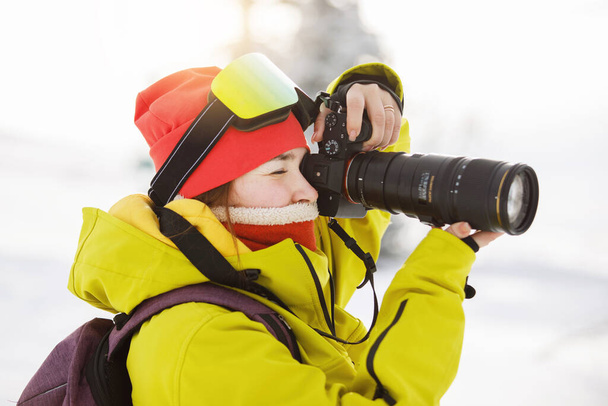 Female photographer in ski equipment takes pictures with a camera with a huge lens. Winter clothes: red pants and a warm yellow jacket, knitted hat, ski goggles. Selective focus. - Φωτογραφία, εικόνα