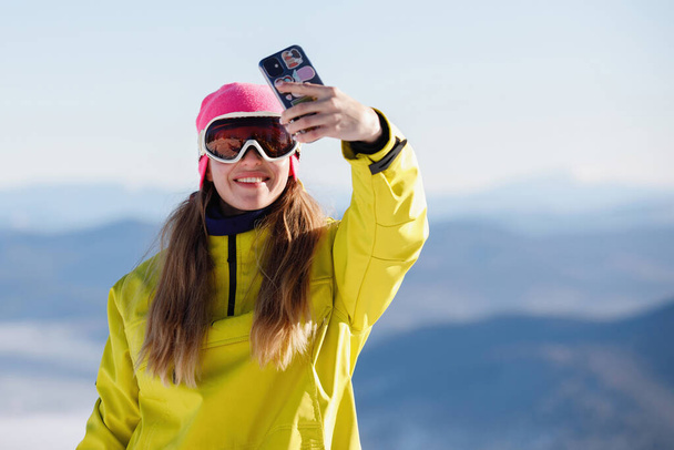 Woman in ski clothes takes a selfie against the backdrop of blue mountains. Yellow or mustard jacket with a hood, a knitted hat, ski goggles. Healthy lifestyle. Sports concept. Selective focus. - Photo, Image