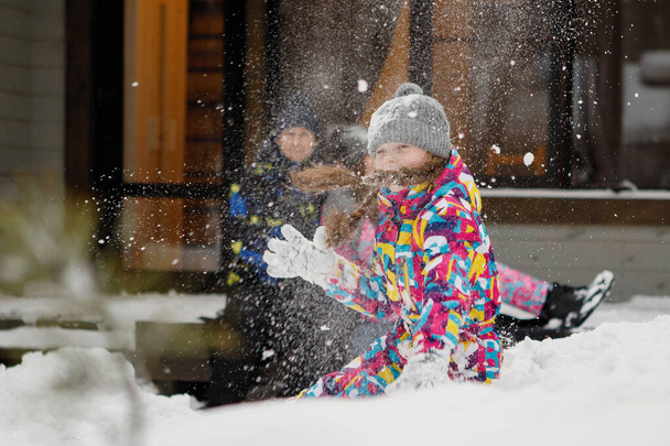 Winterspaß, Freizeit und Familienurlaub Konzept. Mädchen mit Zöpfen wirft Schnee in einer Schneewehe vor ein Fenster zu Hause und Familie? Selektiver Fokus, verschwommener Hintergrund. - Foto, Bild