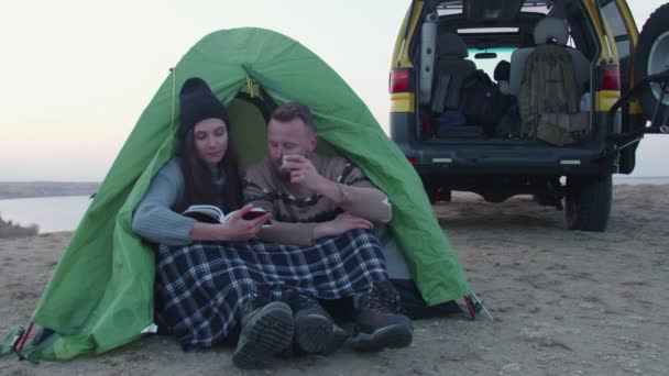 Young man and woman sits near the tent and the car, drinks a hot tea and read the book in the mountain - Footage, Video