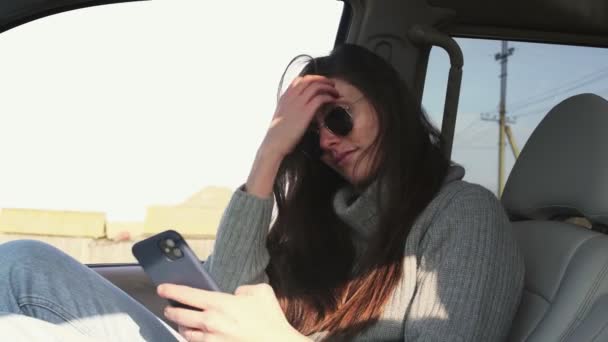 Young woman with sunglasses sits in car and takes selfie in summer morning - Footage, Video