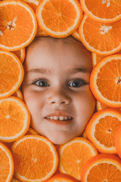 Das Gesicht eines hübschen kleinen Mädchens ist umgeben von geschnittenen Orangen-, Orangen- und Fruchtstücken, die reich an Vitamin C sind.. - Foto, Bild