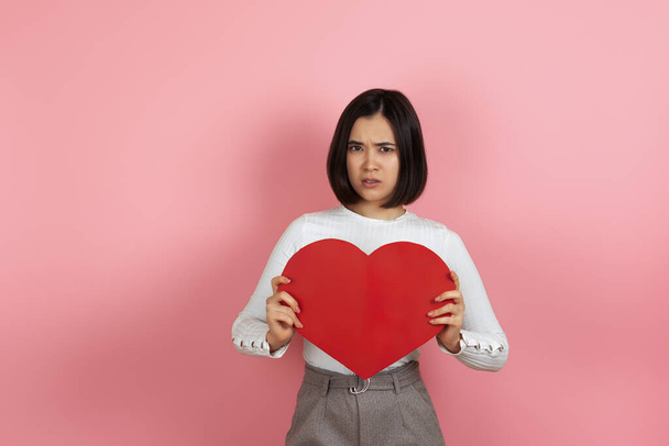 close up sad upset frowning resentful asian woman holding big red paper heart isolated on pink background - 写真・画像