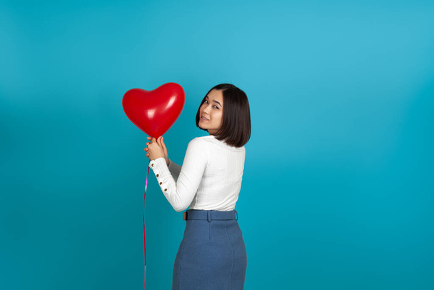 Mulher asiática de volta e segurando um balão vermelho na forma de um coração com duas mãos, isolado no fundo azul claro - Foto, Imagem