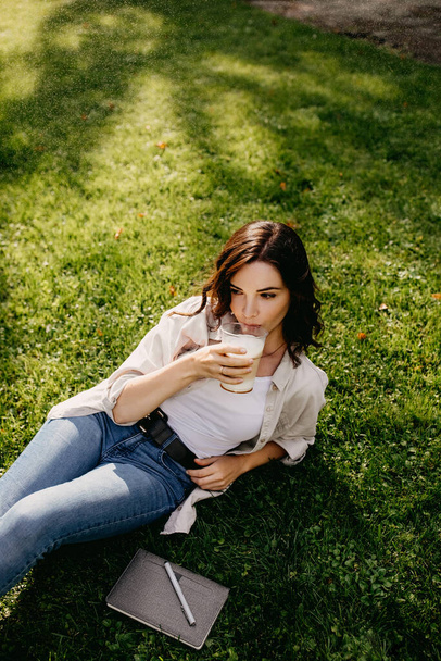 retrato de hermosa mujer joven con taza de plástico de café con leche relajarse en la hierba en el parque - Foto, Imagen