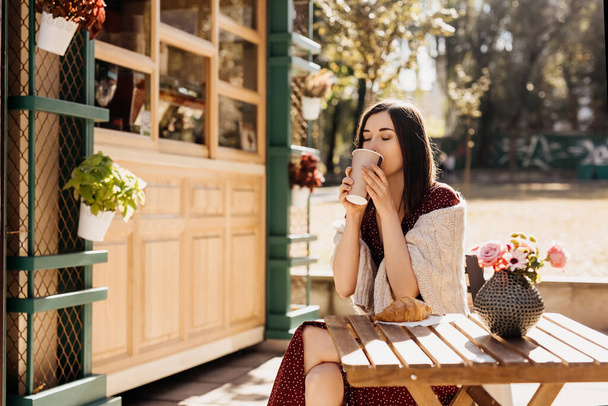 portrait of beautiful young woman sitting in street cafe with paper cup of hot drink and croissant - Fotografie, Obrázek
