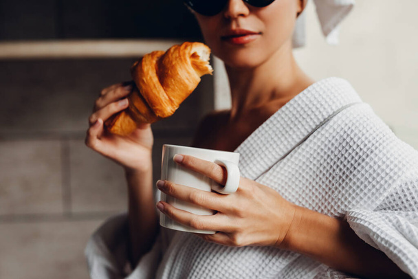 Ayçöreği ve bir fincan kahveyle havluyla ve bornozuyla otelde dinlenen güzel genç bir kadın. - Fotoğraf, Görsel