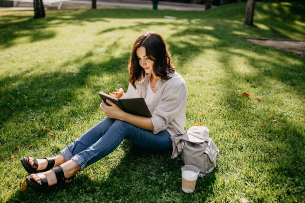 Porträt einer schönen jungen Frau, die im Park auf Gras sitzt und in Notizbuch schreibt - Foto, Bild