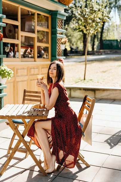 retrato de hermosa joven sentada en la cafetería de la calle con taza de papel de bebida caliente y croissant - Foto, Imagen