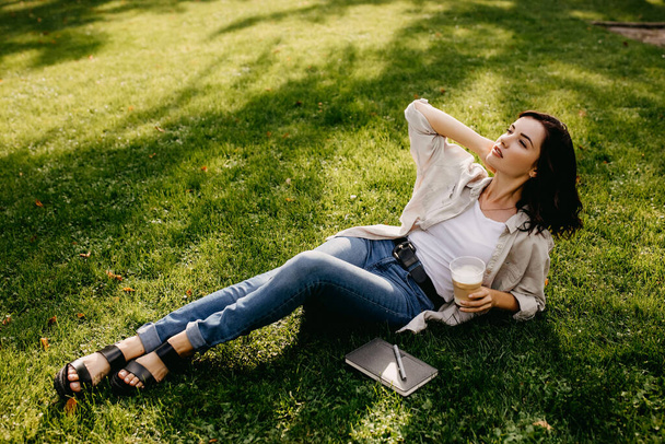 portret pięknej młodej kobiety z plastikową filiżanką latte relaksującej się na trawie w parku - Zdjęcie, obraz