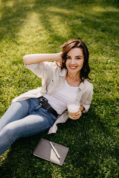 Porträt einer schönen jungen Frau mit Plastikbecher Latte, die es sich auf dem Rasen im Park gemütlich macht - Foto, Bild