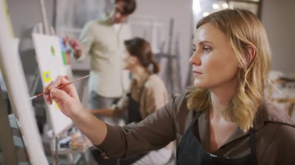 Середня крупним планом концентрована красива жінка, що малює пензлем на полотні, в той час як викладач чоловічих мистецтв розмовляє зі студентом на фоні яскравої сучасної художньої студії
 - Кадри, відео