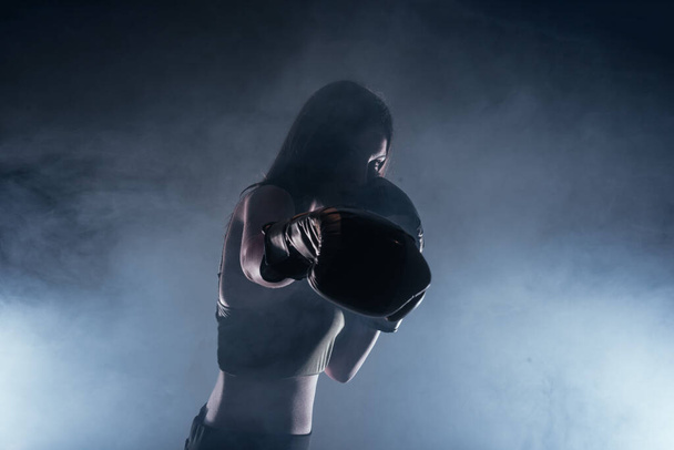 Тёмное контрастное фото с дымом на фоне сфокусированной сильной женщины в боксёрских перчатках, практикующей удары - Фото, изображение