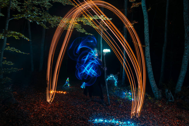 Hechicero (mago) lanzando un hechizo mientras está de pie en los bosques mágicos. Sostiene su bastón mágico. La iluminación mágica. Celebración de vacaciones de otoño. Concepto de misterio y pesadilla - Foto, imagen