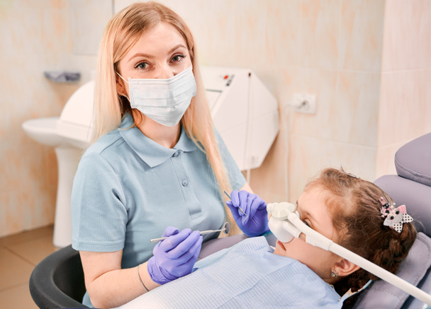 Tıbbî maske takan dişçi, elinde diş aletleriyle dişçi koltuğuna uzanmış diş hekiminde solunum ilacıyla yatıyor. Pediatrik, yatıştırıcı dişçilik ve diş bakımı kavramı. - Fotoğraf, Görsel