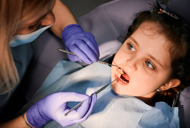Primer plano de la adorable niña con la boca abierta mientras el dentista revisa los dientes de los niños, examinando los dientes de los niños con explorador dental y espejo. Concepto de odontología pediátrica y cuidado dental. - Foto, Imagen