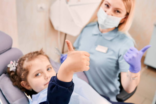 Focus on girl's hand giving thumbs up while woman dentist in medical mask sitting beside kid, showing approval gesture after dental procedure. Concept of pediatric dentistry and dental care approval. - Foto, Bild