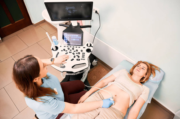 Top view of female patient lying on daybed while having ultrasonography procedure in clinic. Woman sonographer doing abdominal ultrasound scanning for patient. Concept of healthcare and sonography. - Photo, Image