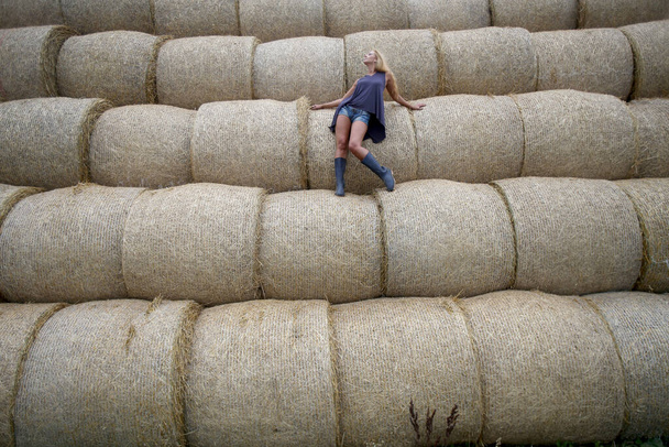 Mujer rubia con botas de goma posando en un campo sobre un rollo de heno - Foto, imagen