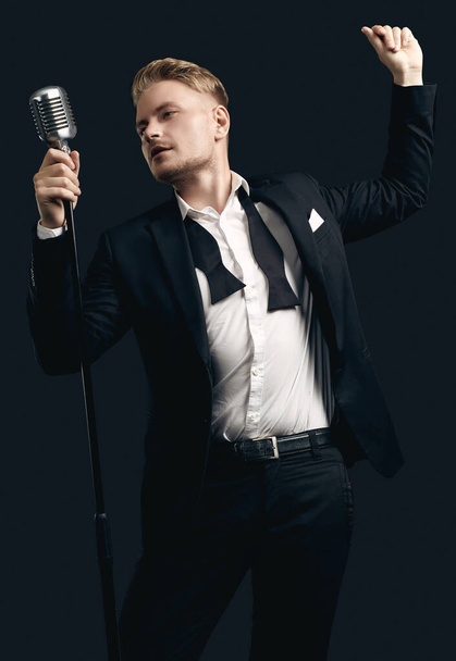 Retrato del guapo cantante hombre rubio en elegante esmoquin y pajarita posando con micrófono vintage sobre fondo de estudio negro - Foto, Imagen