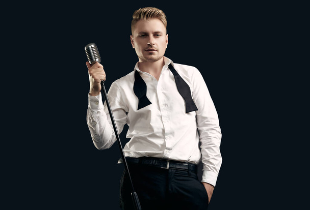 Portrait of handsome blond man singer in elegant tuxedo and bow tie posing with vintage microphone on black studio background - Photo, Image