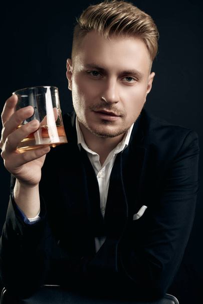Portrait of handsome blond man in elegant tuxedo and bow tie posing with glass of whiskey on black studio background - Photo, Image