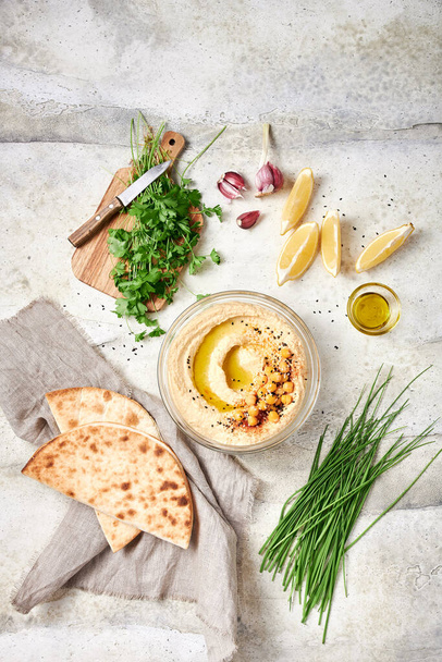 Freshly made hummus with chickpeas, olive oil and paprika in glass bowl on light grey table, with lemon slices, garlic cloves, chive, parsley, flatbreads - Foto, Imagem